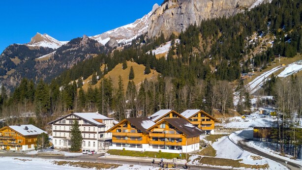 Un conjunto de edificios de madera con balcones, frente a un paisaje de montañas nevadas y cielo azul despejado.