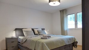 Un dormitorio moderno con una cama tapizada en gris, una mesita de noche con cajones y una ventana con cortinas.