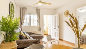 A cozy living room with a sofa, a dining table with plates and bowls, a ceiling fan, and a window with curtains. - Bungalow PINK LAGOON (Torrevieja)