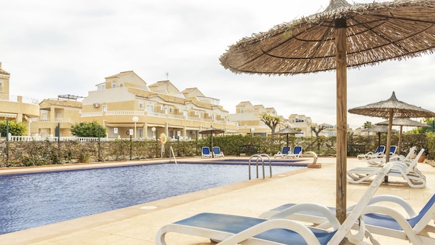 A swimming pool with lounge chairs and umbrellas, surrounded by buildings. - Bungalow PINK LAGOON (Torrevieja)
