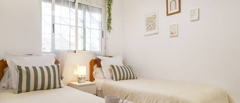 A bedroom with two beds, a bedside table with a lamp, a window with sheer curtains, and wall art.