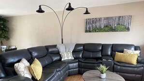 En moderne stue med en sort lædersofa i hjørnesofa-opstilling, et rundt sofabord og et billede af birketræer på væggen.
