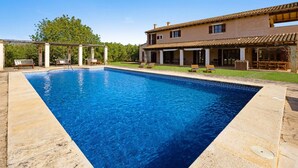 En swimmingpool med et pergolaoverdækket område og liggestole, omgivet af et hus med et tegltag og en overdækket terrasse.