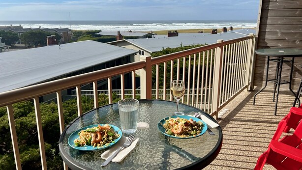 A glass table with two plates of food and a glass of wine on a balcony overlooking the ocean. - Gearhart House G638 (Gearhart)