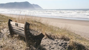 A bench on a sandy beach with a view of the ocean and a distant hill. - Gearhart House G638 (Gearhart)