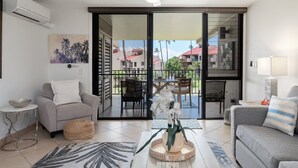 A living room with a sofa, armchair, and a coffee table with a plant. There is a ceiling fan, a sliding glass door leading to an outdoor area with a table and chairs. - Remodeled Condo Across Beach w Pool AC - Kamaole Sands 7307 (Kihei)