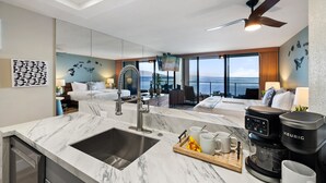Une chambre d’hôtel moderne dotée d’un coin cuisine, d’une grande fenêtre offrant une vue sur l’océan, d’un ventilateur de plafond et d’une machine à café.