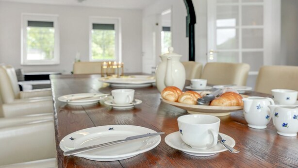 Un coin repas avec une longue table dressée pour le petit-déjeuner, comprenant des croissants, des tasses et des assiettes.