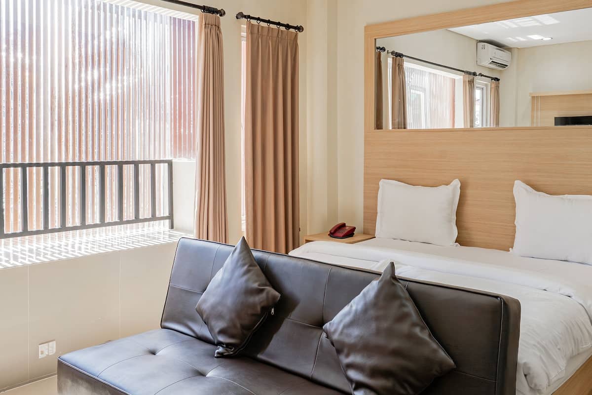 Une chambre d’hôtel avec un lit, une table de chevet avec un téléphone, une fenêtre avec des rideaux et un canapé avec des coussins.