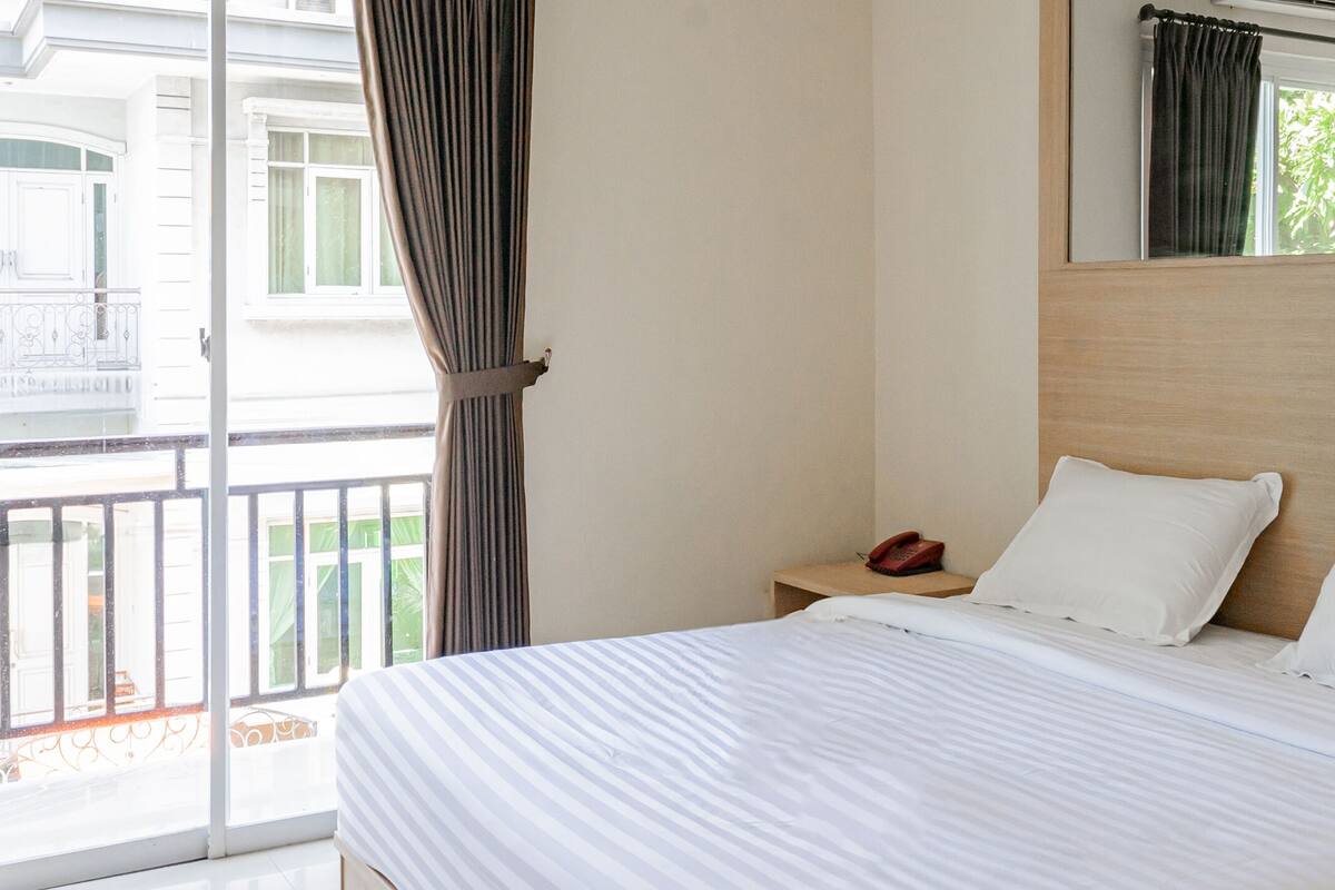 Une chambre d’hôtel avec un lit, une table de chevet avec un téléphone et une vue sur un balcon doté d’une balustrade.