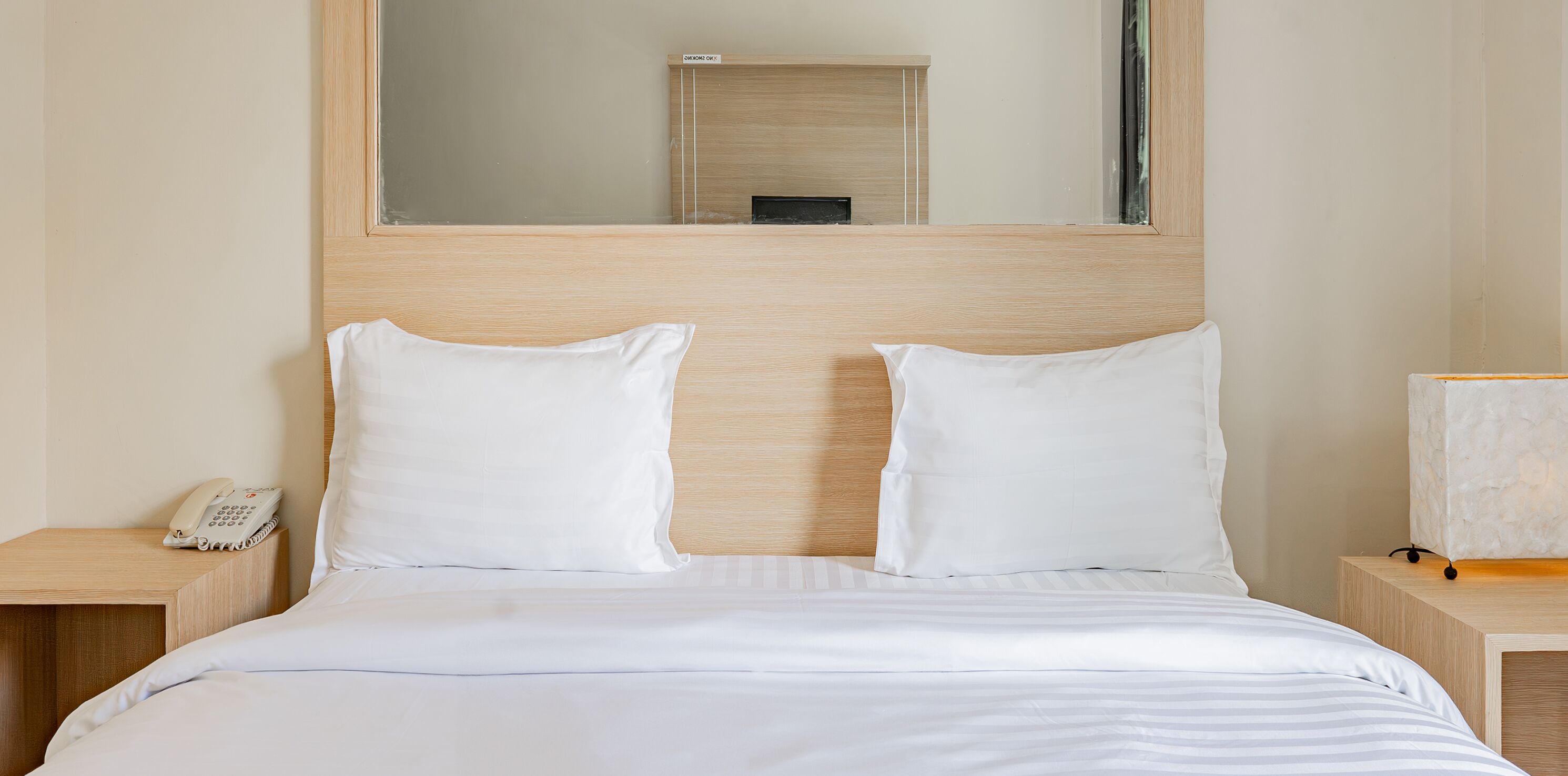 A hotel room with a wooden headboard, two white pillows, a striped bedspread, a bedside table with a phone, and a nightstand with a lamp.