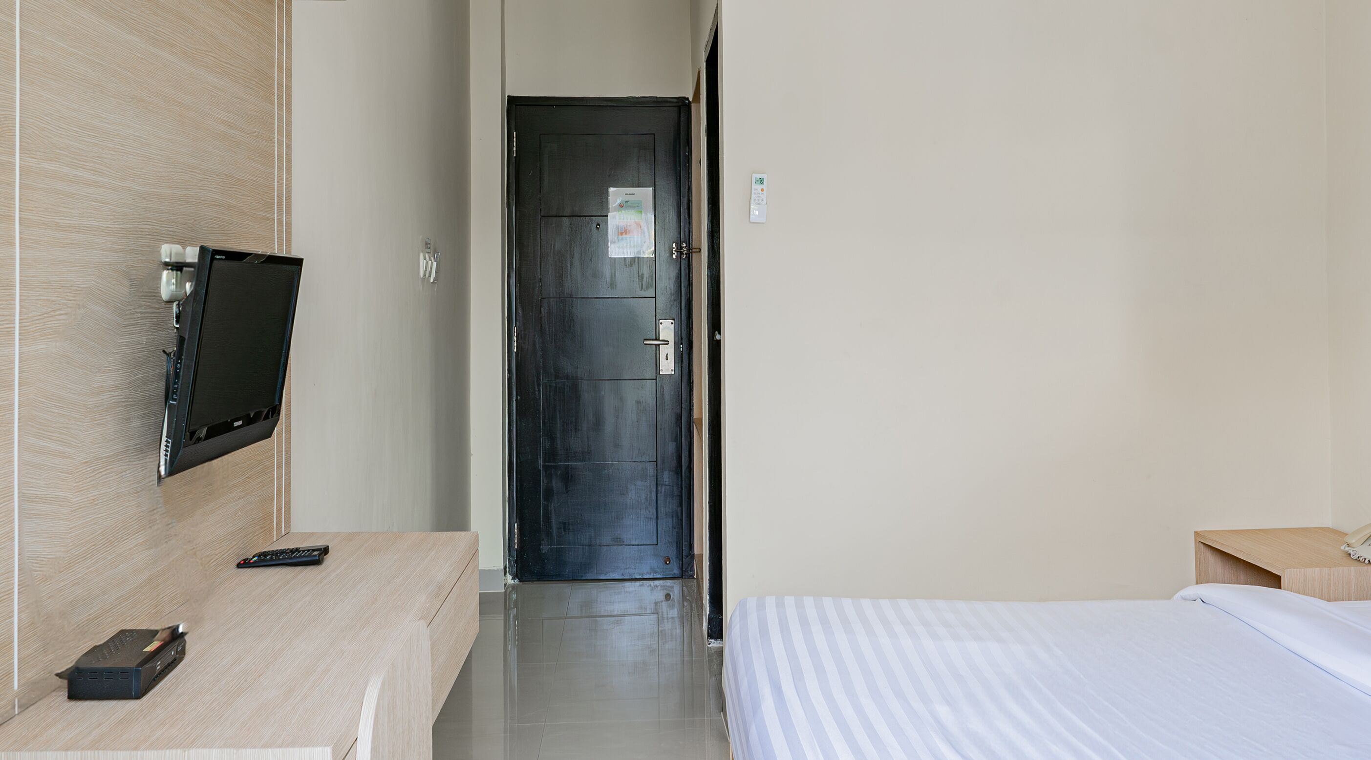 A hotel room with a bed, a bedside table, a TV mounted on the wall, a door, and a light switch.