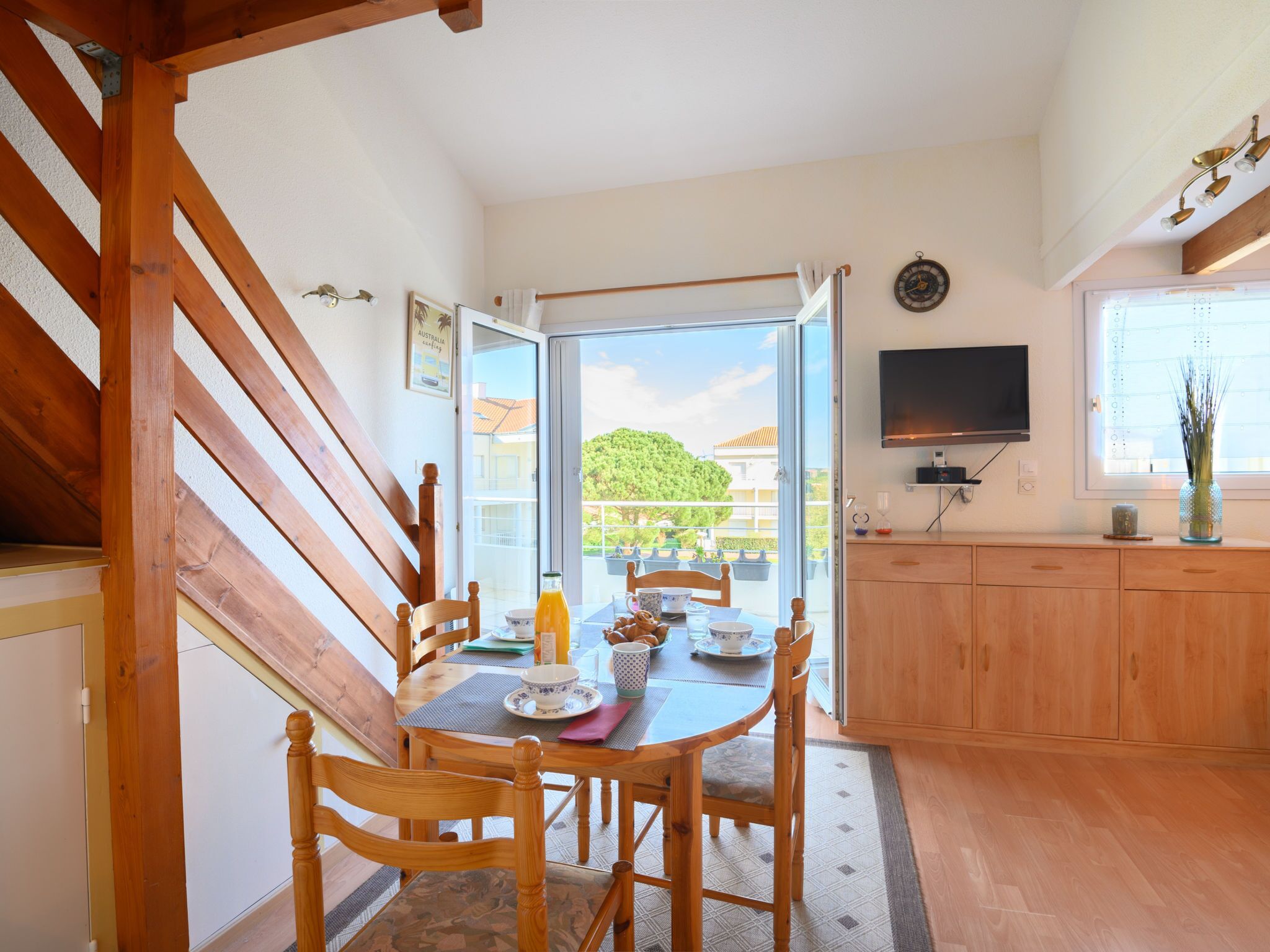 Uma sala de jantar com uma mesa e cadeiras de madeira, uma cozinha com armários de madeira, uma TV fixada na parede e vista para o exterior por uma porta de vidro.