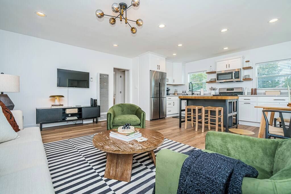 A modern living room with a sofa, a coffee table, and a green armchair. There is a kitchen area with a refrigerator, microwave, and a dining table with stools.