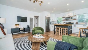 Una sala de estar moderna con un sofá, una mesa de centro y un sillón verde. Hay una zona de cocina con refrigerador, microondas y una mesa de comedor con taburetes.