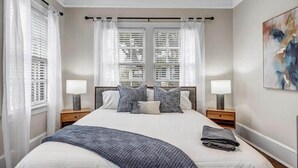A bedroom with a large bed, two bedside tables with lamps, a window with blinds, and a painting on the wall.