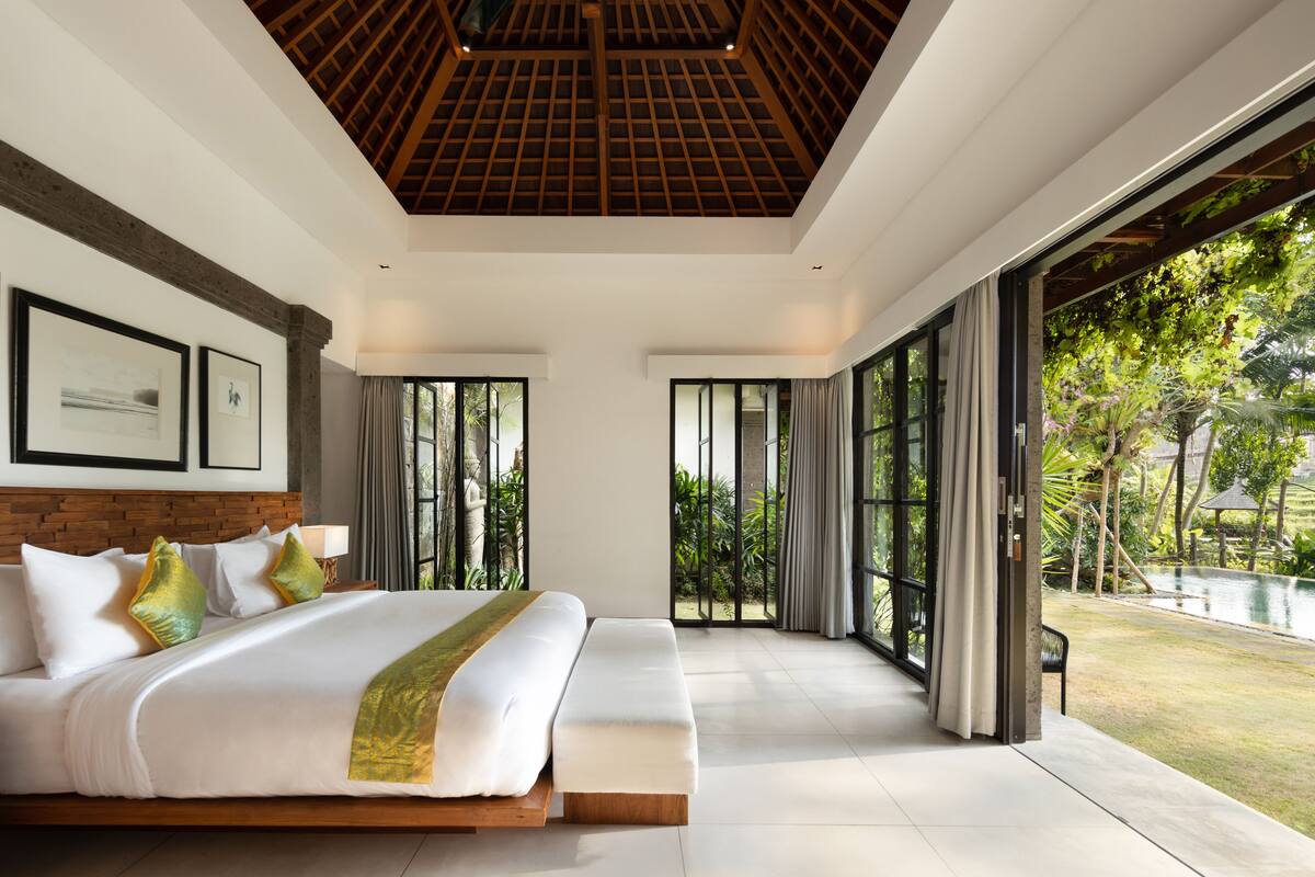 A spacious bedroom with a large bed, a wooden ceiling, and a view of a lush garden and pool.