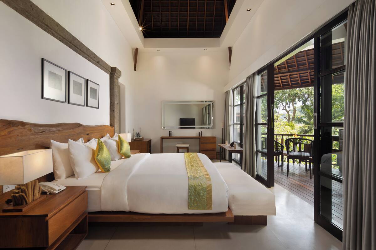 A modern bedroom with a large bed, bedside tables, a desk, and a chair. There is a large window with a view of greenery outside.