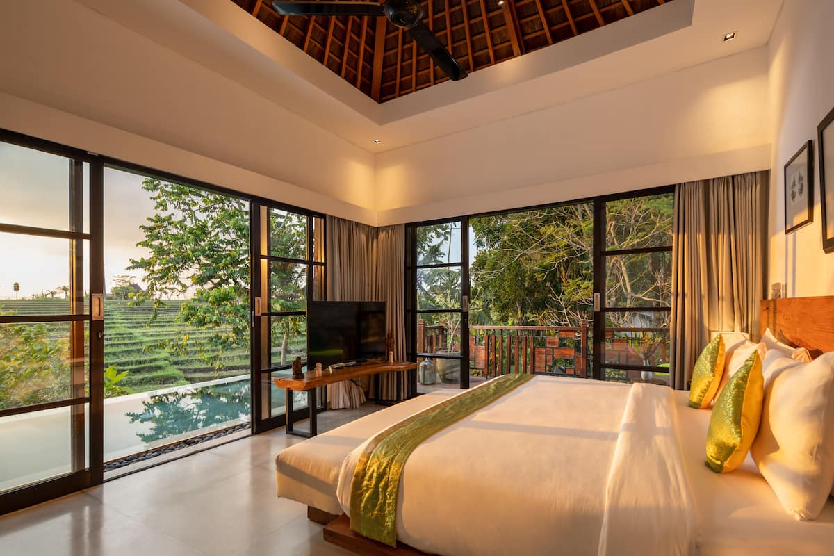 A spacious bedroom with a large bed, a flat-screen TV, and a balcony with a view of a lush garden.