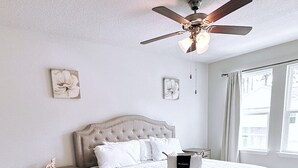 A bedroom with a tufted headboard, a bed with white bedding, a wooden nightstand, a ceiling fan, and a large window with curtains. - New Cozy SanDestin Beach TownHome/30A (Santa Rosa Beach)