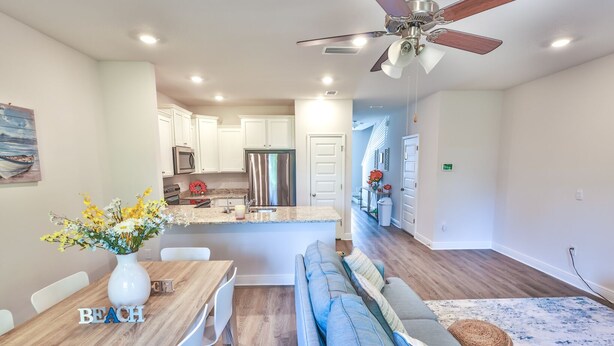A modern kitchen with white cabinets, a stainless steel refrigerator, and a dining area with a wooden table and white chairs. - New Cozy SanDestin Beach TownHome/30A (Santa Rosa Beach)