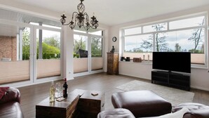 Een ruime woonkamer met een grote televisie, houten meubels en uitzicht door de glazen deuren.