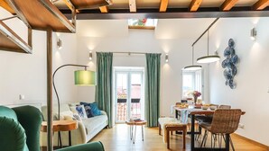 A spacious living area with a sofa, dining table, and chairs. There is a balcony with a view, and the room has wooden flooring and a high ceiling with exposed beams. - Vita Portucale Setubal Deluxe Apartment (Setúbal)
