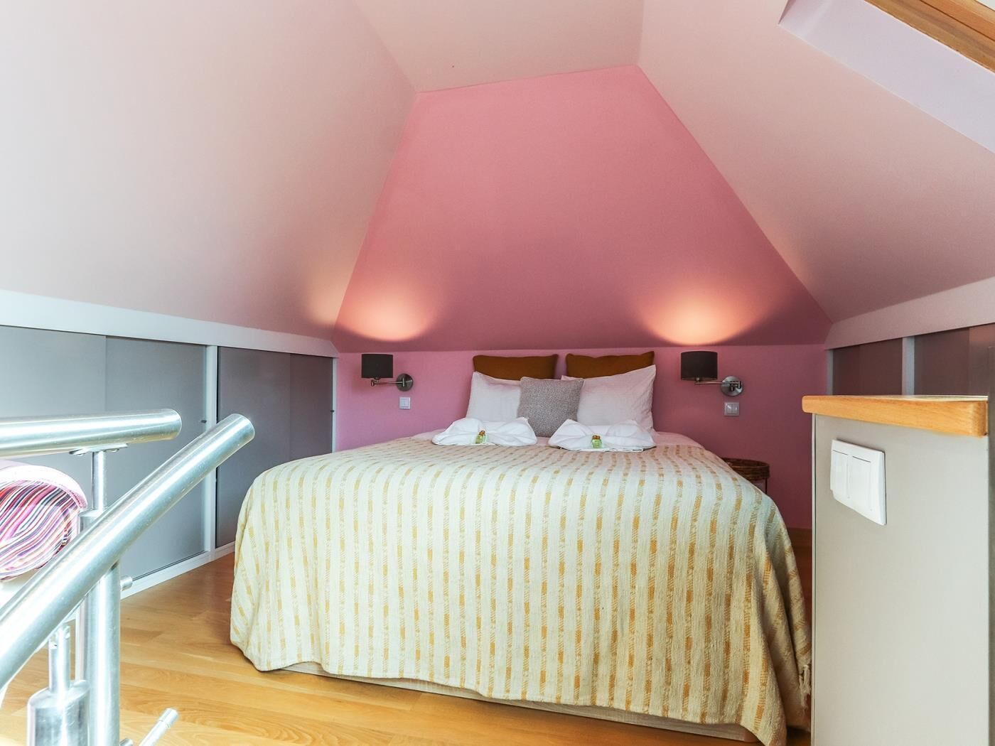 A bedroom with a sloped ceiling, a bed with striped bedding, two wall-mounted lamps, and a wooden railing.
