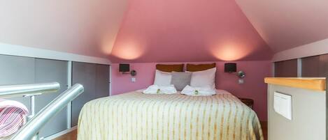 A bedroom with a sloped ceiling, a bed with striped bedding, two wall-mounted lamps, and a wooden railing.