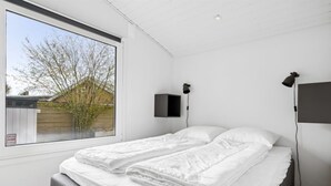 Une chambre avec une grande fenêtre, un lit recouvert de draps blancs, deux haut-parleurs fixés au mur et un panneau d’alarme bien visible.