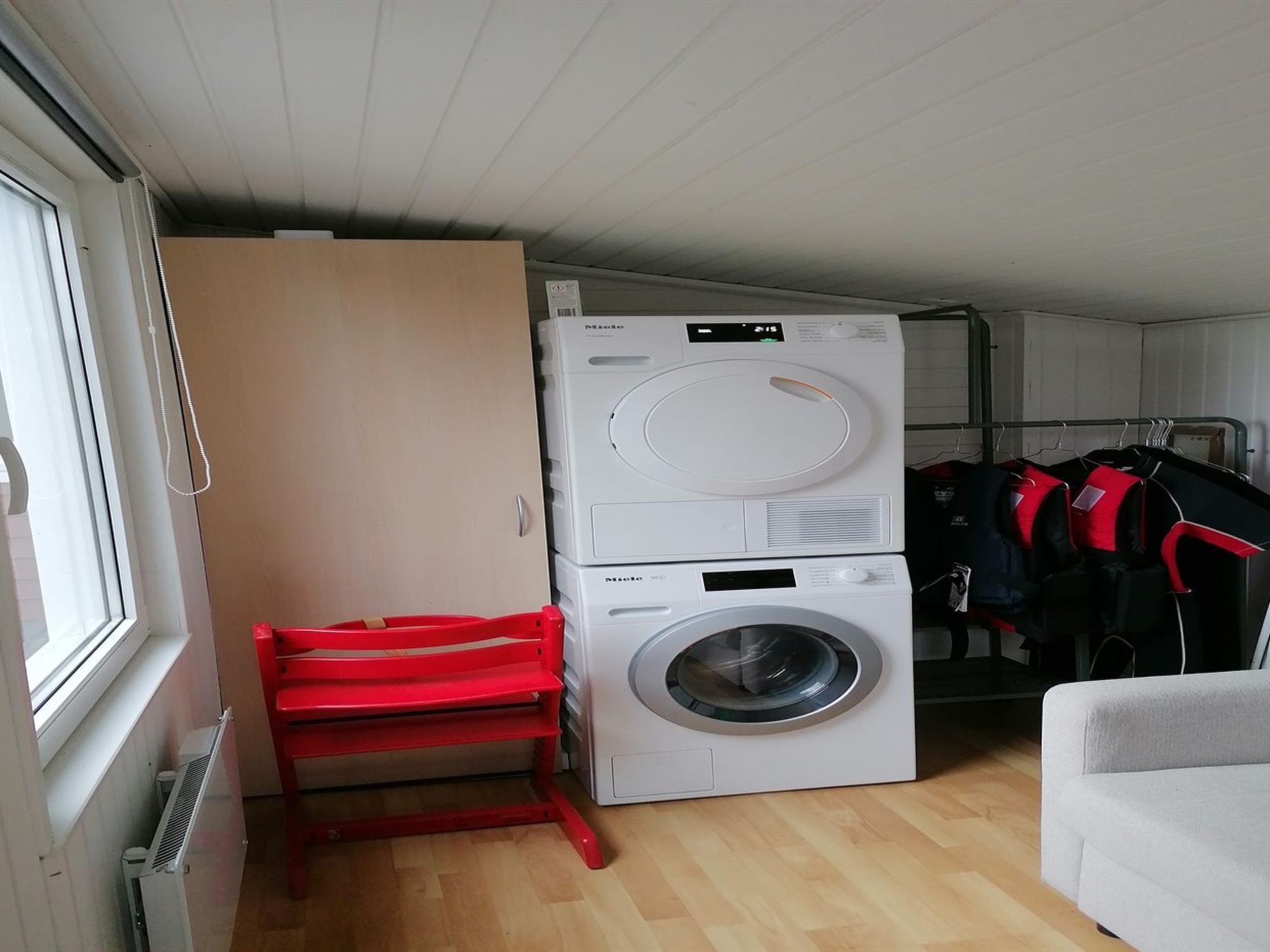 A laundry room with stacked washing machines and dryers, a red folding chair, and a grey sofa.
