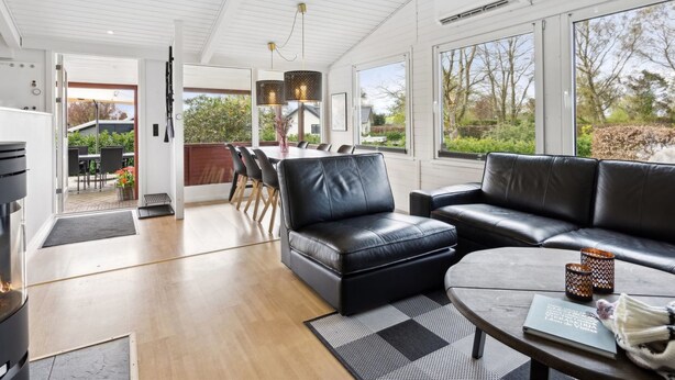 Un salon moderne avec un canapé en cuir noir, un coin repas comprenant une table et des chaises, et de grandes fenêtres offrant une vue sur l’extérieur.