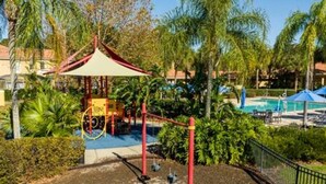 Un terrain de jeux avec une structure rouge et jaune, entouré de palmiers et d’un espace piscine.