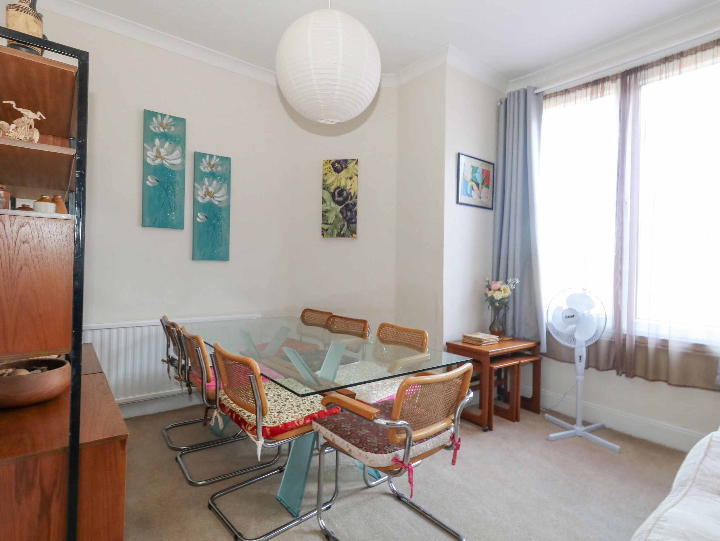 A dining area with a glass table surrounded by chairs, a white ceiling fan, and artwork on the walls.