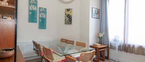 A dining area with a glass table surrounded by chairs, a white ceiling fan, and artwork on the walls.