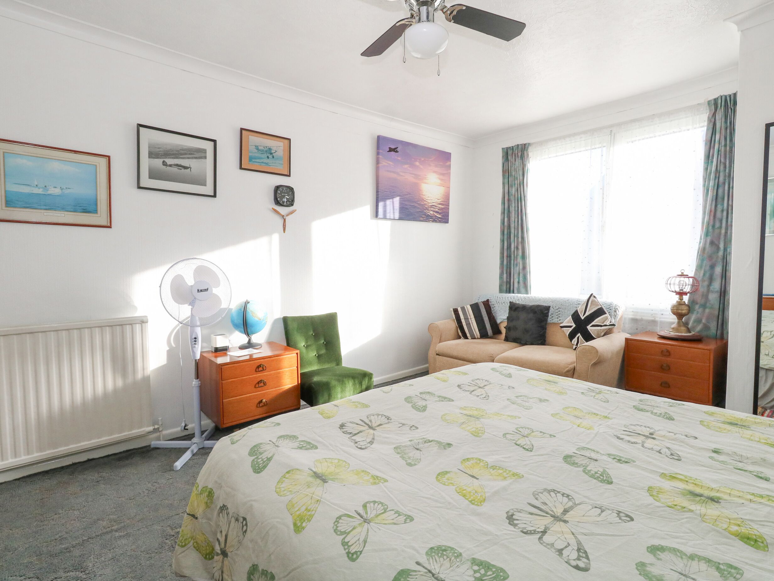 A bedroom with a bed, a green armchair, a sofa, a dresser, a globe, a fan, and a window with curtains.