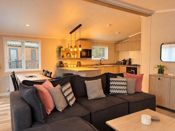 A modern living room with a sectional sofa, a dining table, and a kitchen area with wooden cabinets and a microwave.
