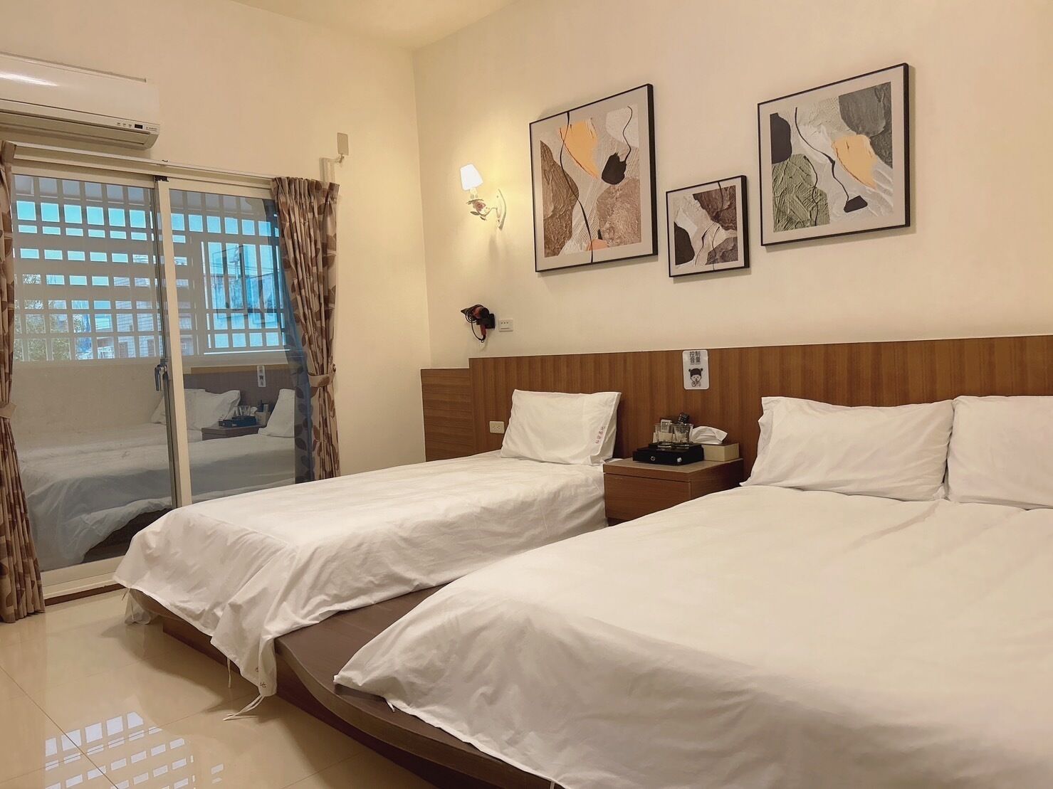 Habitación de hotel con dos camas, una puerta corrediza de vidrio, una lámpara de pared y arte abstracto.