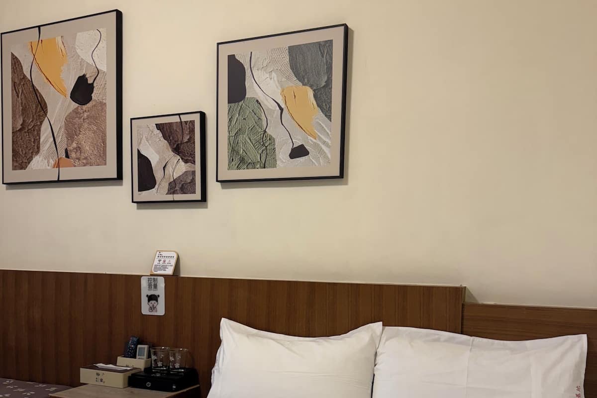 A hotel room with a bed, a wooden headboard, a nightstand with a lamp, and three abstract paintings on the wall.