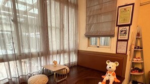 A cozy room with a wooden bench, a small table with a vase, and a teddy bear. There are curtains, a window with blinds, and a shelf with items. - Secret base B&B (Taitung)