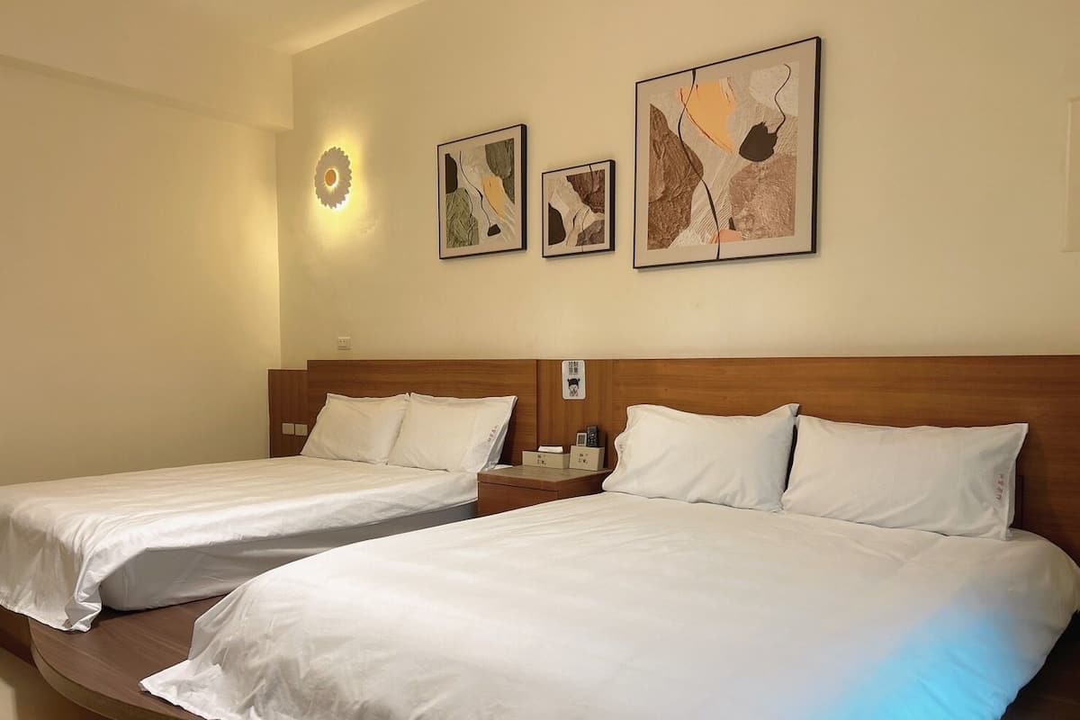 A hotel room with two single beds, a wooden headboard, a nightstand with a lamp, and three framed artworks on the wall.