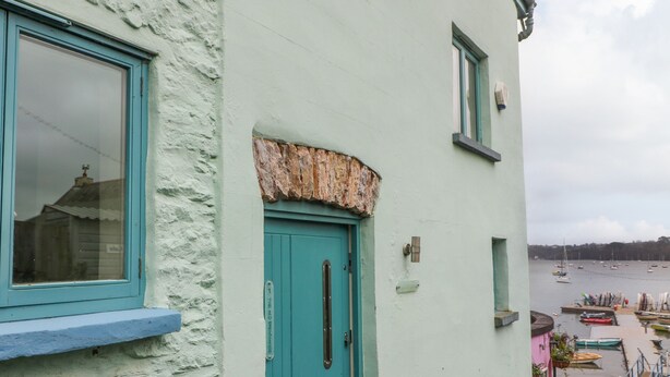 A coastal building with a turquoise door and windows, a weathered stone ledge above the door, and a view of boats and water in the background.
