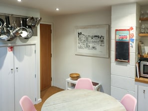 A kitchen with white cabinets, a wooden door, a framed black and white cityscape, a small white table with a bowl, and pink chairs.