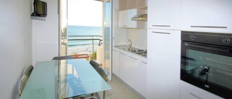A compact kitchen with a glass table and chairs, a built-in oven, and a view of the ocean through an open door.
