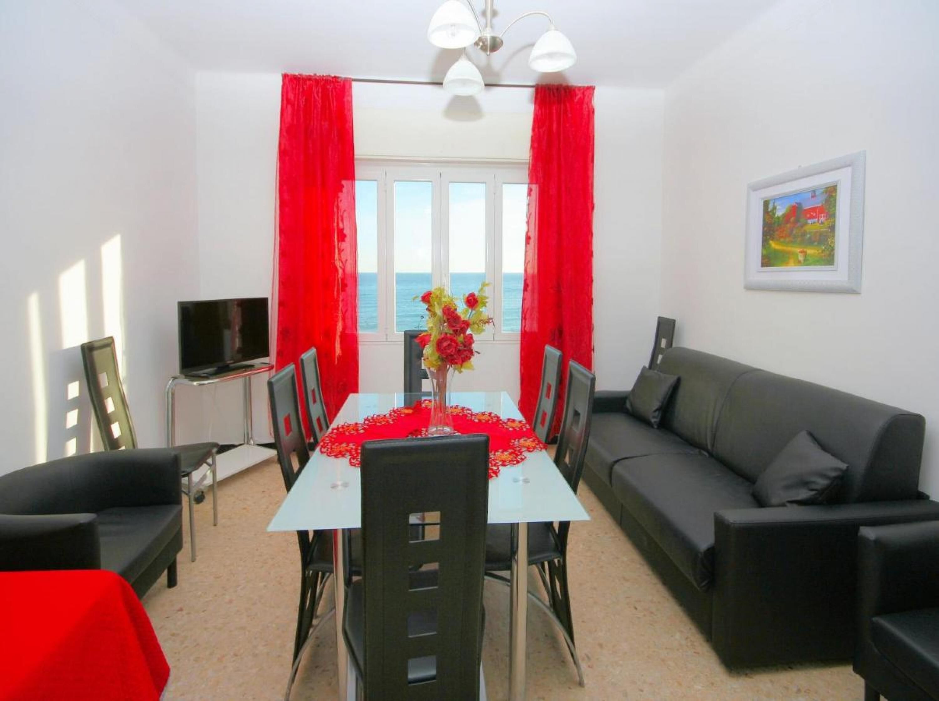 A room with a dining table set for four, a grey sofa, a TV, and a window with red curtains.
