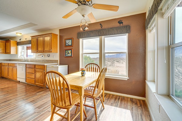 A kitchen with wooden cabinets, a dining table with chairs, a ceiling fan, and a large window with a view of the mountains. - Sky Islands Retreat in Rodeo w/ Mountain Views! (Rodeo)
