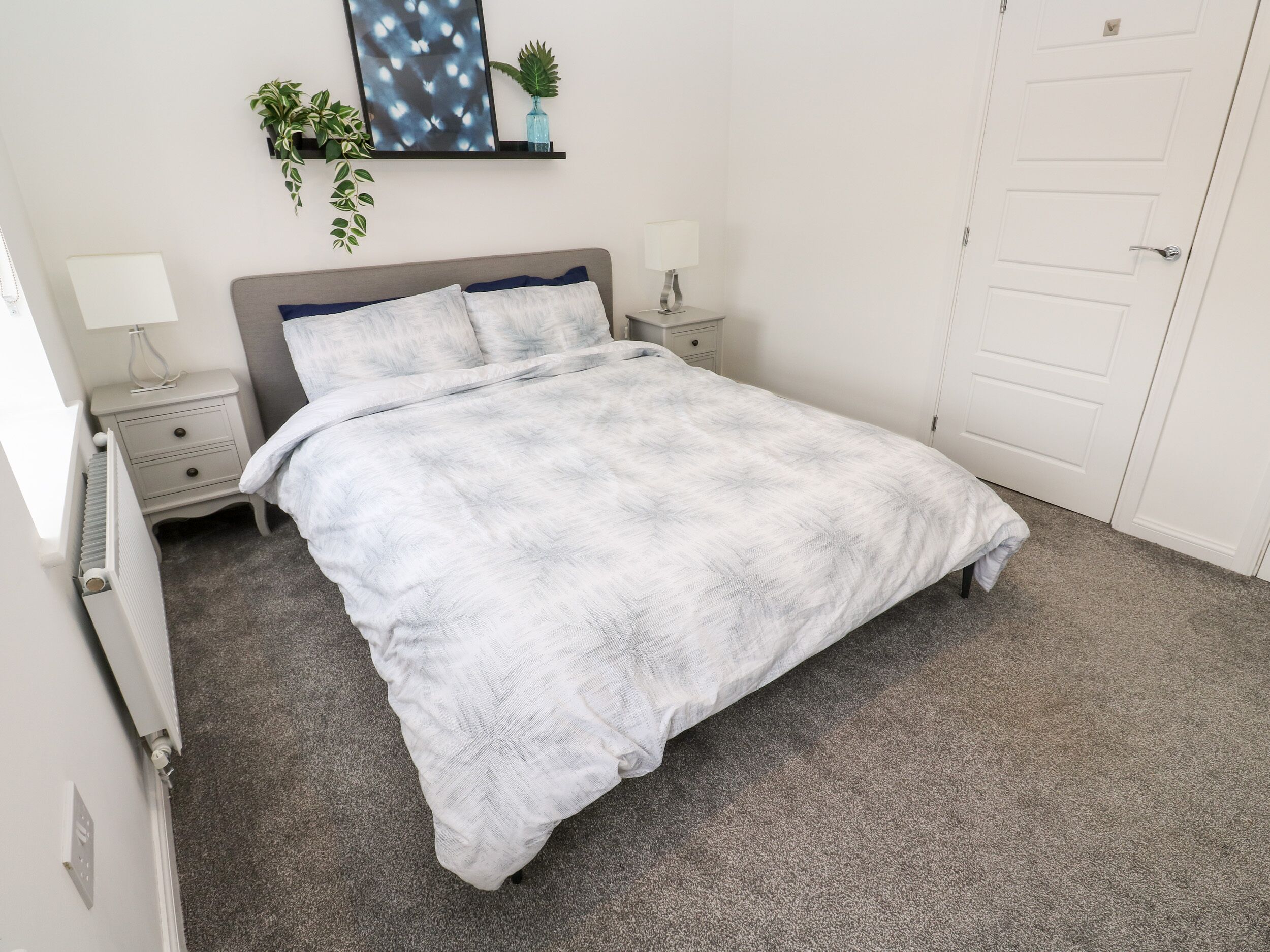 A neatly made bed with a grey headboard, a bedside table with a lamp, and a wall-mounted shelf with plants.