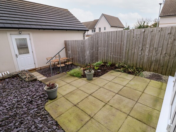 A residential backyard with a paved patio, a wooden fence, a door, and a small garden area with potted plants.