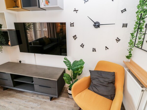 A living room with a large wall clock, a flat-screen TV, a sofa, and a potted plant. - 20 Bishops Way (Falmouth)