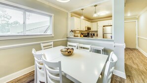 A dining area with a white table and chairs, a basket of bread, and a view of the kitchen in the background.